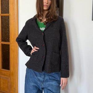 May include: A dark gray blazer with red buttons and two front pockets. The blazer is worn over a green t-shirt. The person is wearing blue jeans. The blazer is a classic style, suitable for various occasions.