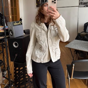 May include: A light beige jacket with multiple pockets and a button-up front. The jacket is paired with black pants and a gray beanie with the letters "AD" on it. The person is holding a phone in front of their face.