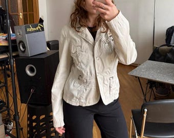 May include: A light beige jacket with multiple pockets and a button-up front. The jacket is paired with black pants and a gray beanie with the letters "AD" on it. The person is holding a phone in front of their face.