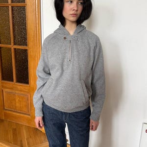 May include: A gray hooded sweatshirt with a quarter-zip closure and a front pocket. The sweatshirt is paired with dark blue denim jeans. The person is standing in front of a wooden door and a framed picture.
