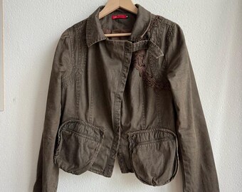 May include: A brown jacket with two large front pockets and decorative embroidery on the collar and chest. The jacket is hanging on a wooden hanger against a white wall. The jacket has a button closure.