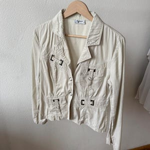 May include: A light beige jacket with a collared neckline and button closure. The jacket features two chest pockets and two lower pockets, each with a flap and a decorative buckle. The jacket is hanging on a wooden hanger.