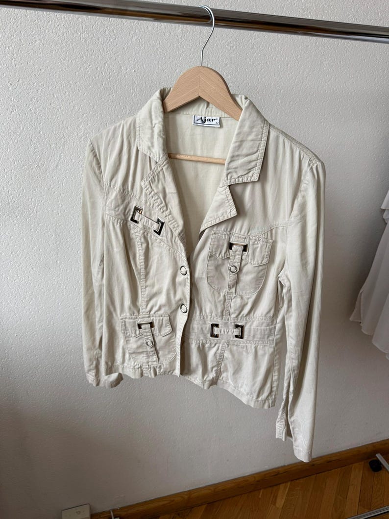 May include: A light beige jacket with a collared neckline and button closure. The jacket features two chest pockets and two lower pockets, each with a flap and a decorative buckle. The jacket is hanging on a wooden hanger.