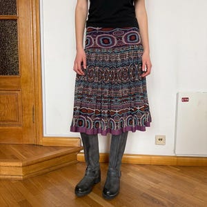 May include: A patterned skirt with a purple hemline, paired with gray boots. The skirt features a repeating geometric design in shades of blue, brown, and purple. The top is a solid black color.