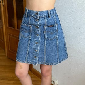 Puede incluir: Falda vaquera vintage en un lavado azul claro. La falda presenta una botonadura frontal, dos bolsillos delanteros y una pequeña etiqueta con el logo. La falda tiene una silueta clásica en forma de A.