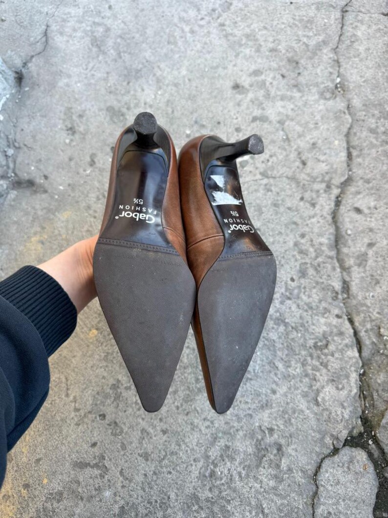 May include: A pair of brown leather high heels with pointed toes and stiletto heels. The soles are dark brown, and the heels are black. The brand name "Gabor Fashion" is printed on the insole. The shoes are size 5 1/2.