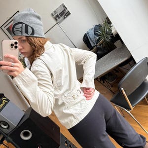 May include: A light beige jacket with long sleeves and a button-up front. The jacket has a tailored fit and features a small pocket detail on the back. The person is also wearing a gray beanie with a black logo and black pants.