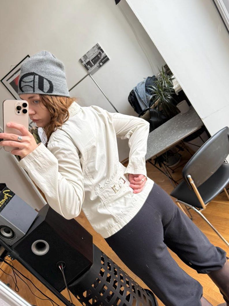 May include: A light beige jacket with long sleeves and a button-up front. The jacket has a tailored fit and features a small pocket detail on the back. The person is also wearing a gray beanie with a black logo and black pants.