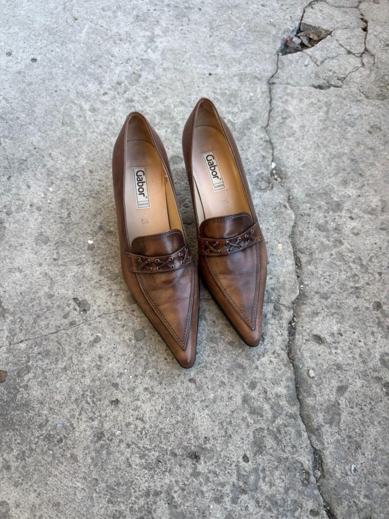 May include: A pair of brown leather Gabor court shoes with pointed toes. The shoes have a low heel and a decorative strap. The Gabor logo is visible inside the shoe.