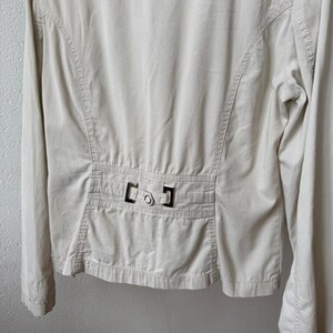 May include: A light beige jacket with long sleeves. The back of the jacket features a tailored design with vertical seams and a decorative buckle detail at the waist. The jacket is made of a smooth, lightweight fabric.