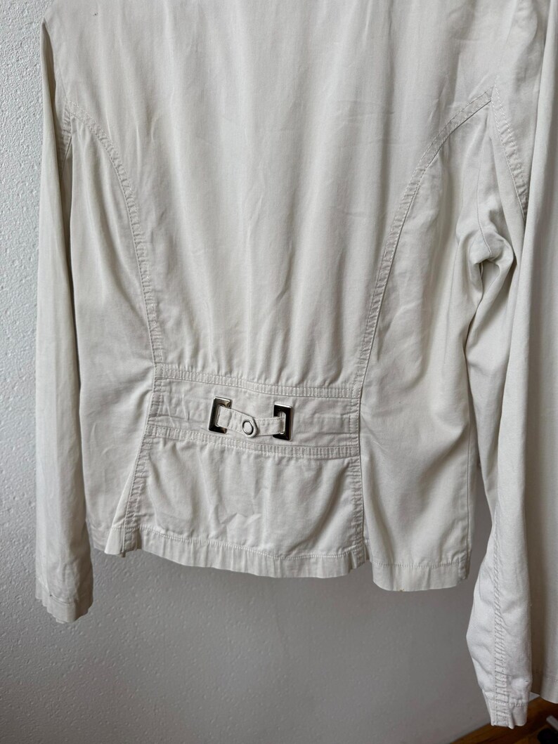 May include: A light beige jacket with long sleeves. The back of the jacket features a tailored design with vertical seams and a decorative buckle detail at the waist. The jacket is made of a smooth, lightweight fabric.