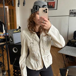 May include: Beige jacket with multiple pockets and metal accents. The jacket is buttoned up and has a tailored fit. The person is wearing a gray beanie with black lettering and black pants. The jacket appears to be made of a lightweight fabric.