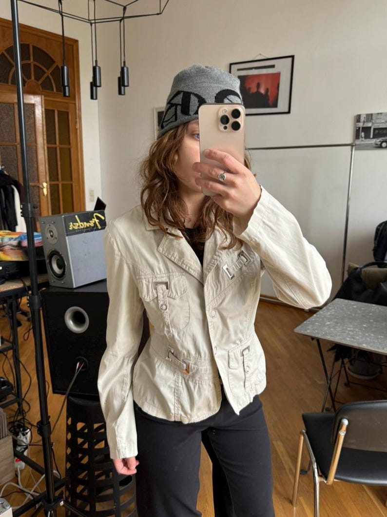 May include: Beige jacket with multiple pockets and metal accents. The jacket is buttoned up and has a tailored fit. The person is wearing a gray beanie with black lettering and black pants. The jacket appears to be made of a lightweight fabric.