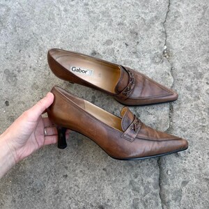 May include: A pair of brown leather heeled loafers. The shoes feature a pointed toe, decorative stitching, and a small heel. The brand name "Gabor" is visible inside one shoe. The shoes are on a concrete surface.
