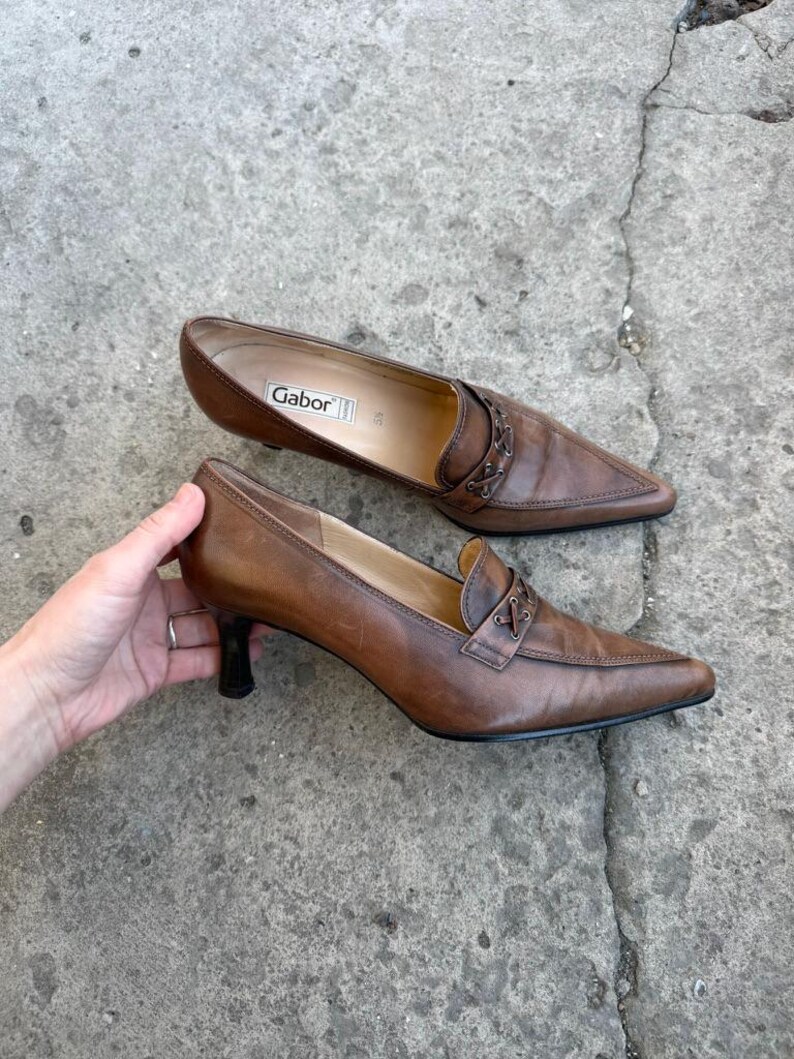 May include: A pair of brown leather heeled loafers. The shoes feature a pointed toe, decorative stitching, and a small heel. The brand name "Gabor" is visible inside one shoe. The shoes are on a concrete surface.
