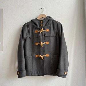 May include: A gray duffel coat with a hood, wooden toggles, and leather patches. The coat is hanging on a wooden hanger against a white wall. The coat has two front pockets and buttoned cuffs.