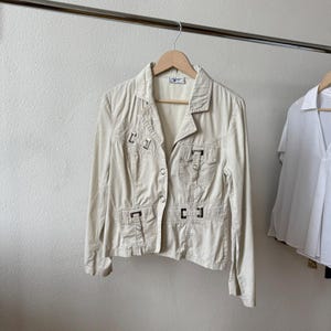 May include: A beige jacket with a collared neckline and button closure, hanging on a wooden hanger. The jacket features multiple pockets with decorative metal accents. The garment is displayed against a neutral background, showcasing its design and details.