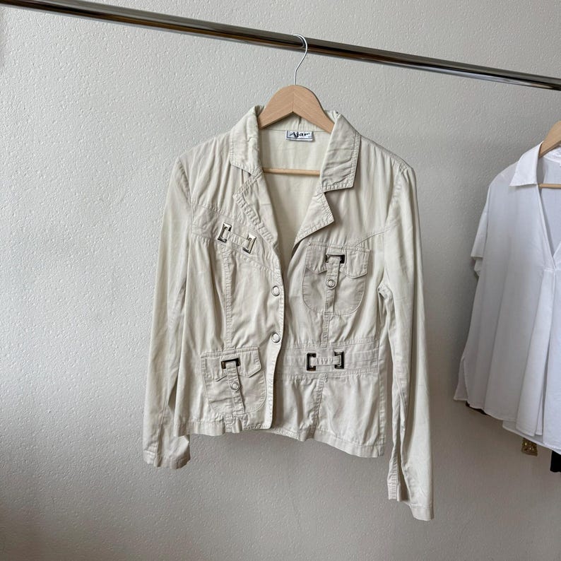 May include: A beige jacket with a collared neckline and button closure, hanging on a wooden hanger. The jacket features multiple pockets with decorative metal accents. The garment is displayed against a neutral background, showcasing its design and details.