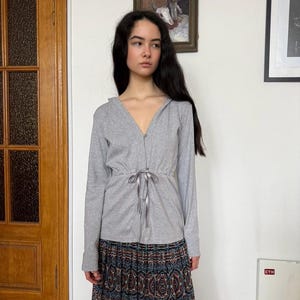 May include: A light gray hooded top with a drawstring waist and long sleeves. The top is paired with a long, patterned skirt in shades of blue, brown, and red. The outfit is worn indoors.