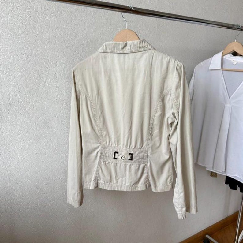 May include: A beige jacket with long sleeves and a collar hangs on a wooden hanger. The back of the jacket features a decorative belt detail with a metal buckle. A white shirt hangs to the right.