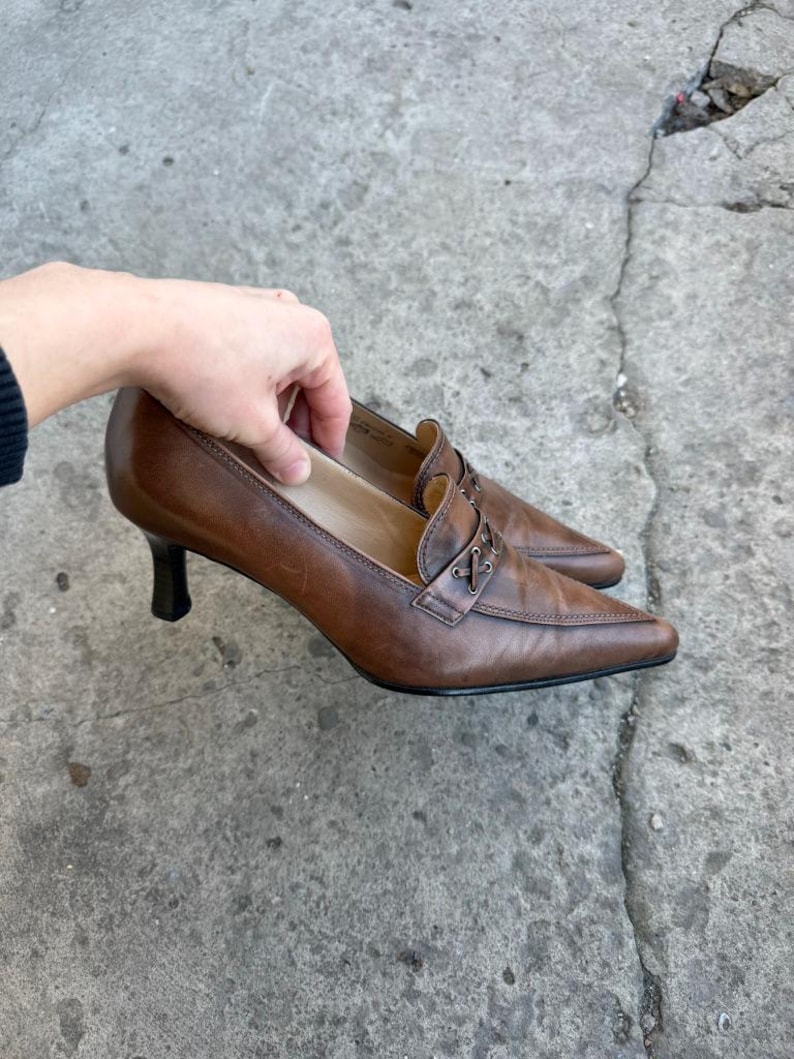 May include: Brown leather heeled shoes with pointed toes. The shoes have a decorative strap across the top and a small, black heel. The shoes are displayed on a concrete surface.