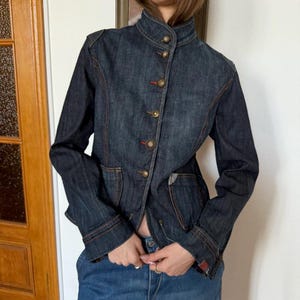 May include: A dark wash denim jacket with a mandarin collar and long sleeves. The jacket features gold-colored buttons with red accents and two front pockets. The seams are highlighted with orange stitching. The model is wearing blue jeans.