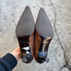May include: A pair of brown leather Gabor Fashion heels, size 5 1/2, with pointed toes and small heels. The soles are dark brown, and the heels are black. The shoes are held up, showing the soles and heels.