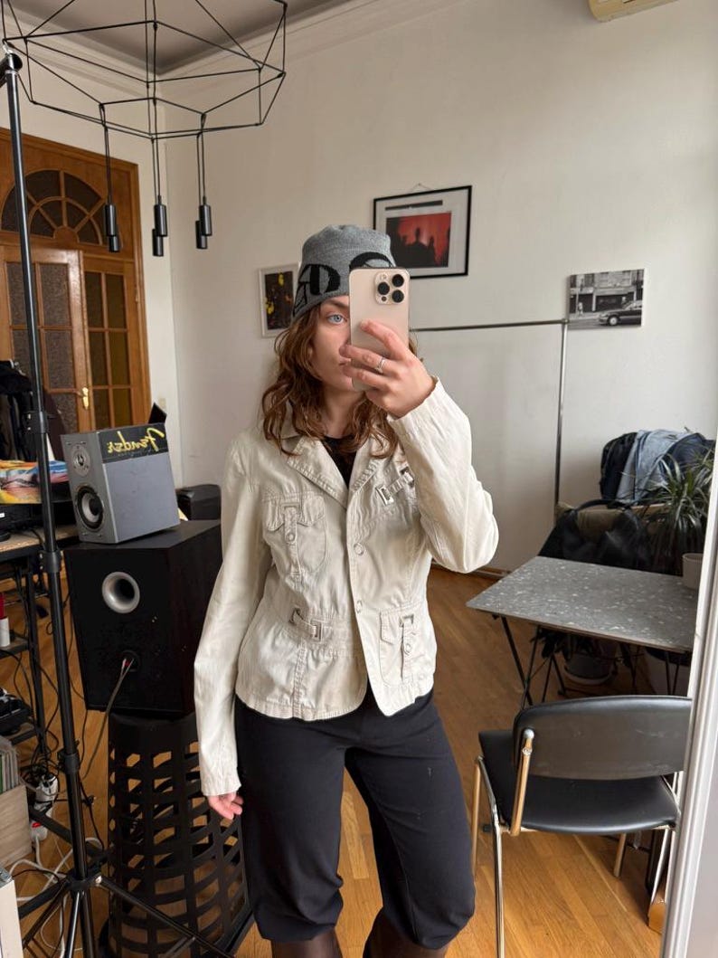 May include: A light beige jacket with multiple pockets and a button-up front. The jacket is paired with black pants and a gray beanie with the letters "AD" on it. The person is holding a phone in front of their face.