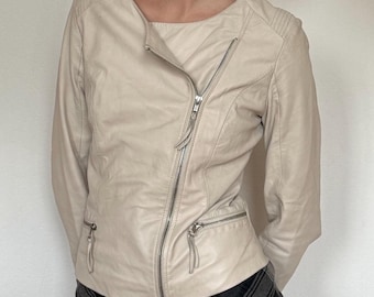 Puede incluir: Chaqueta de cuero color crema con cierre de cremallera diagonal y dos bolsillos con cremallera. La chaqueta tiene cuello redondo y mangas largas. Los hombros tienen un diseño acolchado. La chaqueta se combina con una falda a cuadros negros y grises.