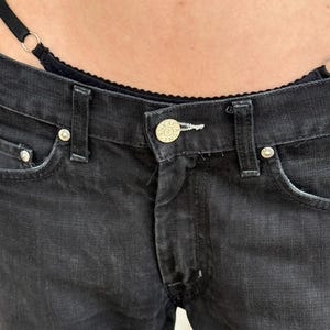 以下が含まれることがあります： ボタン留めとポケットのシルバーリベットが付いた黒いデニムジーンズのクローズアップ。ジーンズはクラシックな5ポケットデザインで、色あせた外観です。ボタンには「JEANS」の文字が刻印されています。