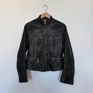 以下が含まれることがあります： 黒い革のジャケット。ジッパー開閉、4つのジッパーポケット、白いステッチのディテール。 ジャケットは白い壁を背景に、木製のハンガーに掛けられています。