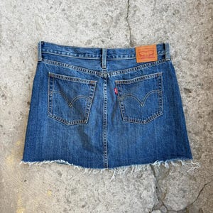 May include: A blue denim skirt with a raw hem. The skirt has two back pockets and a small red tag. A brown leather patch with the brand name is visible at the waistband.