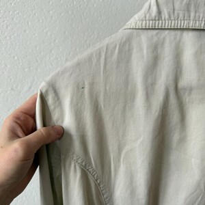May include: Close-up of a light beige collared shirt. The shirt has a button-down design and a tailored fit. The fabric appears to be a lightweight material, suitable for warmer weather. The collar is neatly pressed, and the stitching is visible.