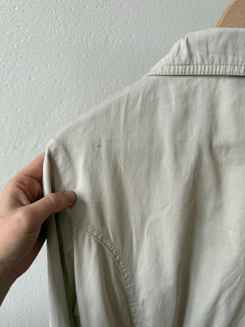 May include: Close-up of a light beige collared shirt. The shirt has a button-down design and a tailored fit. The fabric appears to be a lightweight material, suitable for warmer weather. The collar is neatly pressed, and the stitching is visible.