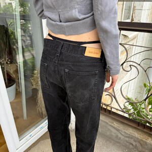 以下が含まれることがあります： 色あせた外観の黒いデニムジーンズ。クラシックな5ポケットデザインで、「Acne Jeans」と書かれた革のタグが付いています。ジーンズはグレーのジャケットと組み合わせています。画像は屋外で撮影されています。