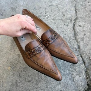 May include: A pair of brown leather loafers with pointed toes. The shoes have a decorative stitched band across the top and are set against a grey concrete surface. The shoes are a size 8.