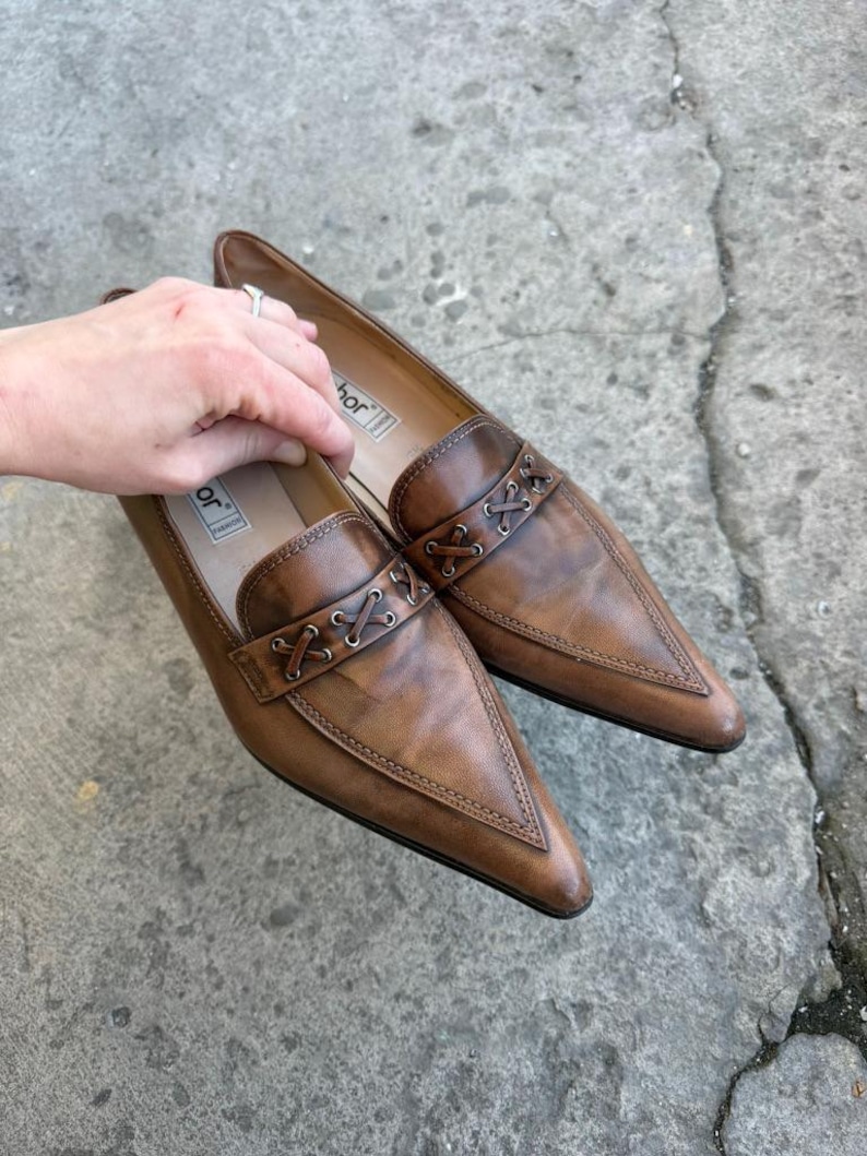 May include: A pair of brown leather loafers with pointed toes. The shoes have a decorative stitched band across the top and are set against a grey concrete surface. The shoes are a size 8.