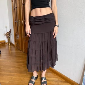 Op de afbeelding: Een donkerbruine rok met ruches en een gerimpelde taille. De rok wordt gecombineerd met een zwarte top en zilveren sandalen. De rok heeft een vloeiend, gelaagd ontwerp, geschikt voor casual of semi-formele gelegenheden.
