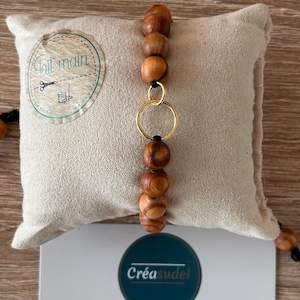 Puede incluir: Una pulsera de cuentas de madera con un anillo dorado y un cordón negro. La pulsera se exhibe sobre un cojín beige. La imagen también muestra una tarjeta de visita con el texto "Créasudel" y una dirección web.