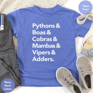 Puede incluir: Una camiseta azul con texto blanco que enumera diferentes tipos de serpientes: Pitones, Boas, Cobras, Mambas, Víboras y Áspides. La camiseta se combina con pantalones y zapatos grises. Texto adicional en la imagen dice "Many More Colors" y "Ships Fast!"