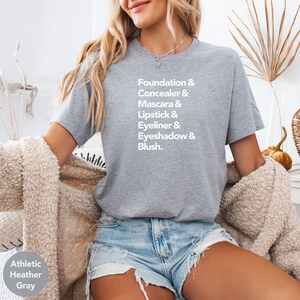 Pode incluir: T-shirt cinzento desportivo com o texto branco "Foundation & Concealer & Mascara & Lipstick & Eyeliner & Eyeshadow & Blush". A modelo usa calções de ganga e um cardigan creme.