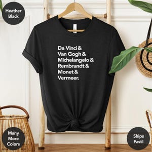May include: Heather black t-shirt with the names of famous artists printed in white: Da Vinci, Van Gogh, Michelangelo, Rembrandt, Monet, and Vermeer. The shirt is displayed on a wooden hanger.