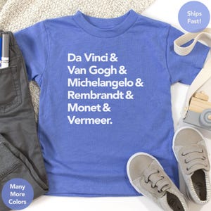 Puede incluir: Una camiseta azul bígaro con los nombres de artistas famosos impresos en blanco: Da Vinci, Van Gogh, Miguel Ángel, Rembrandt, Monet y Vermeer. La camiseta se muestra con pantalones grises y zapatillas de deporte grises claras.