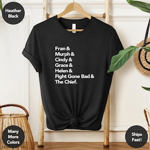 May include: Heather black t-shirt with white text: Fran & Murph & Cindy & Grace & Helen & Fight Gone Bad & The Chief. The shirt is displayed on a wooden rack with a neutral background. Additional text reads "Many More Colors" and "Ships Fast!"