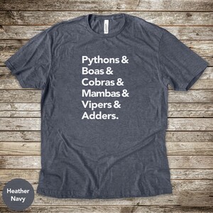 Puede incluir: Camiseta azul marino jaspeado con texto blanco que enumera varios tipos de serpientes: Pitones, Boas, Cobras, Mambas, Víboras y Áspides. La camiseta tiene cuello redondo y es de tejido suave.
