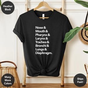 May include: Black heather t-shirt with white text listing parts of the respiratory system: Nose, Mouth, Pharynx, Larynx, Trachea, Bronchi, Lungs, and Diaphragm. The shirt is displayed on a wooden hanger.