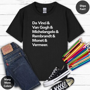 May include: Black t-shirt with white text listing famous artists: Da Vinci, Van Gogh, Michelangelo, Rembrandt, Monet, and Vermeer. The image also shows blue jeans, black sneakers, and colored pencils. The shirt is a Gildan brand.