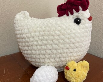 May include: A handmade white crochet chicken with a red comb and black eyes. Beside the chicken are a white crochet egg and a yellow crochet chick with a red beak. The items are on a brown surface.