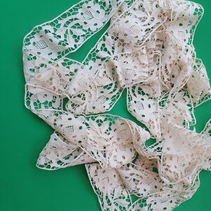 Op de afbeelding: Crèmekleurig kanten lint met een bloemmotief, gepresenteerd tegen een effen groene achtergrond. Het kant heeft een delicaat, opengewerkt patroon, ideaal voor knutselen of versiering.