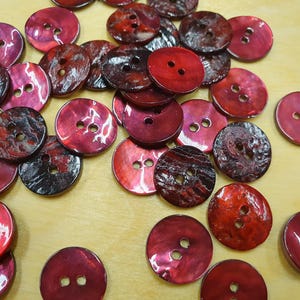 Könnte beinhalten: Eine Sammlung roter und kastanienbrauner Knöpfe mit jeweils zwei Löchern. Die Knöpfe haben eine glänzende Oberfläche und unterschiedliche Muster, einige mit Marmoreffekt. Die Knöpfe sind auf einer hellen Oberfläche verstreut.